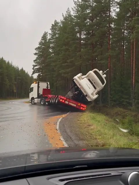 Valkoinen superjahti kallistuu rekan lavetilta metsän reunaan liikenneympyrässä sateisena päivänä.