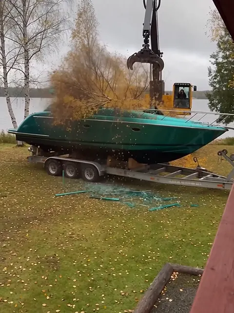 Kaadettu koivu makaa emeraldinvihreän jahdin päällä mökin nurmella, taustalla harvesteri ja järvi.