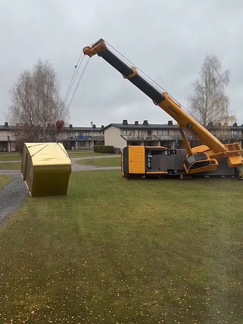 Keltainen autonosturi on kaatunut kyljelleen rivitalon nurmelle, ja kultainen sauna on pudonnut maahan.